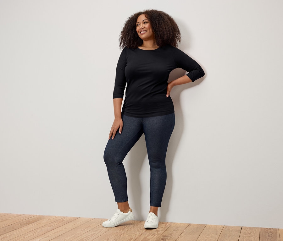 Usmívající se žena pózuje v černém tričku s tříčtvrtečními rukávy, tmavě modrých džeginách a bílých teniskách.