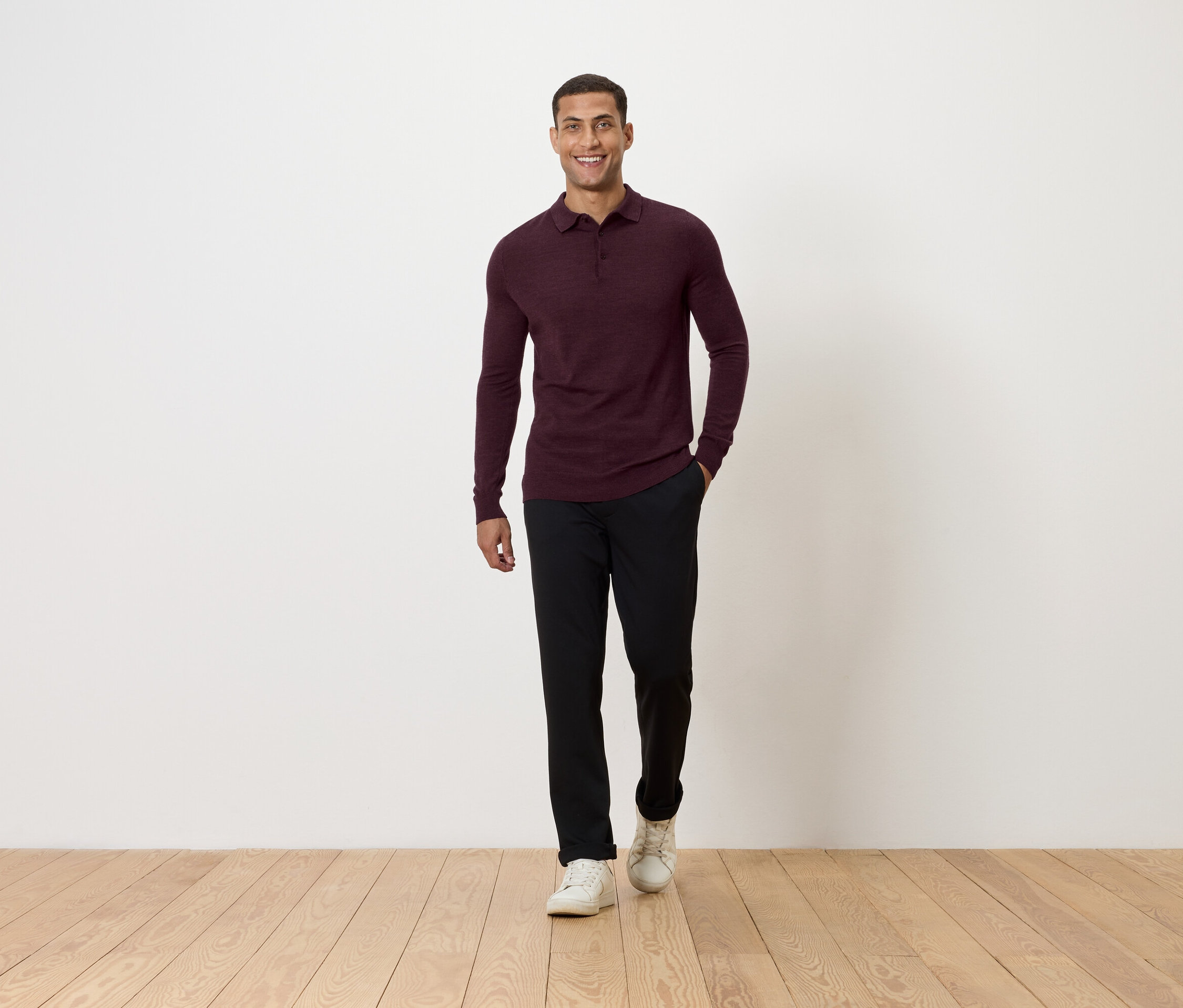 Muž jde po dřevěné podlaze. Má na sobě vínové polo triko s dlouhým rukávem, černé kalhoty a bílé tenisky.