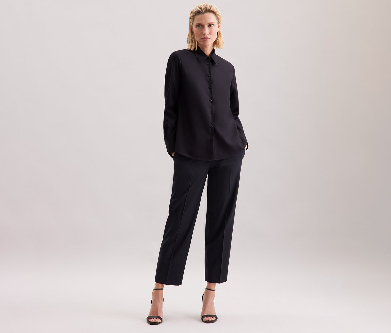 Blond žena stojí na světlém pozadí v černé halence Seidensticker, černých kalhotách a černých sandálech.