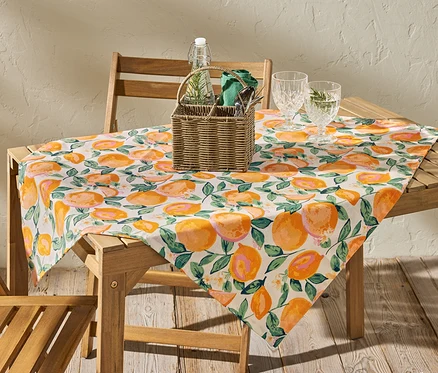 Na dřevěném stole leží omyvatelný ubrus s potiskem oranžových citrusů, cca 90 x 90 cm.