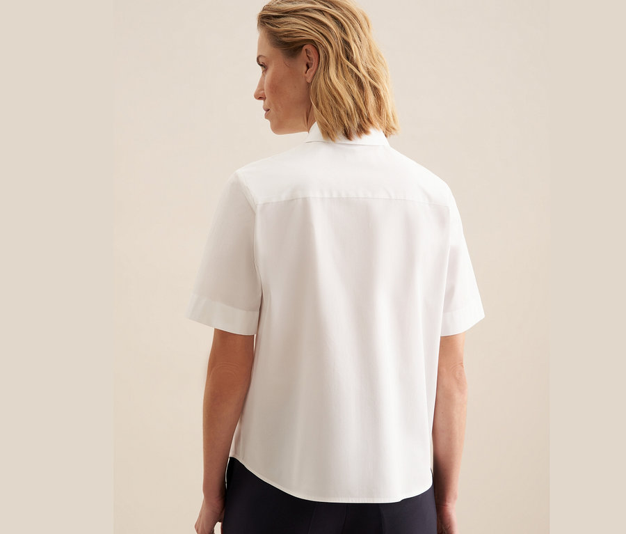 Žena zády k fotoaparátu má na sobě bílou halenku s krátkým rukávem Seidensticker.