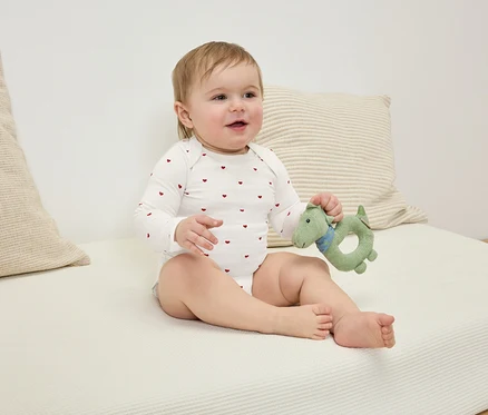 Sedící kojenec v body s červenými srdíčky drží zelenou hračku ve tvaru dinosaura.