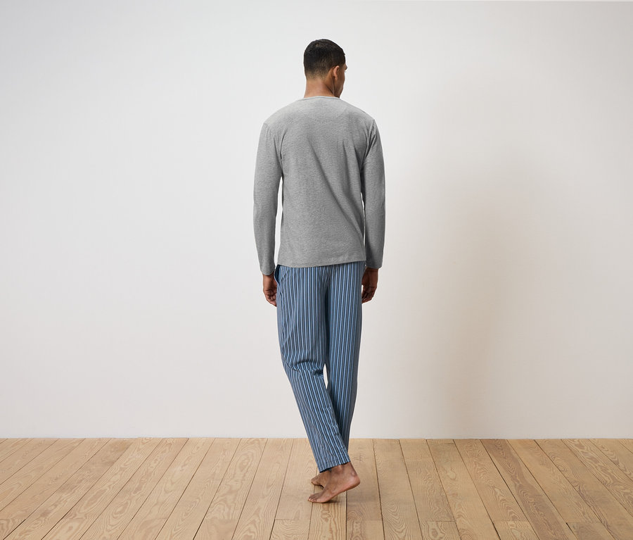 Muž zády k fotoaparátu stojí na dřevěné podlaze. Má na sobě šedé tričko s dlouhým rukávem a modré pruhované pyžamové kalhoty.