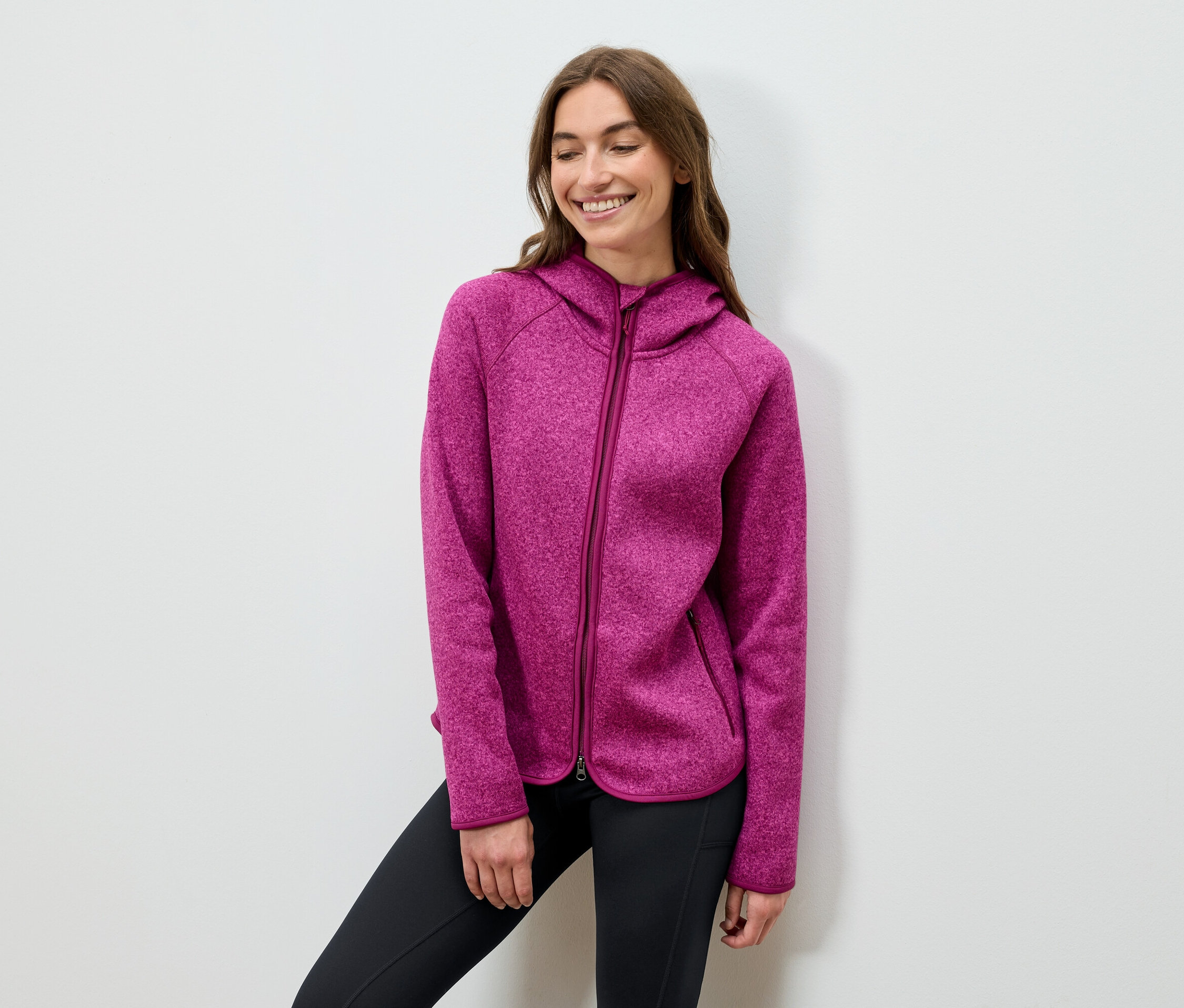 Usmívající se žena pózuje v růžové mikině s kapucí a černých kalhotách.