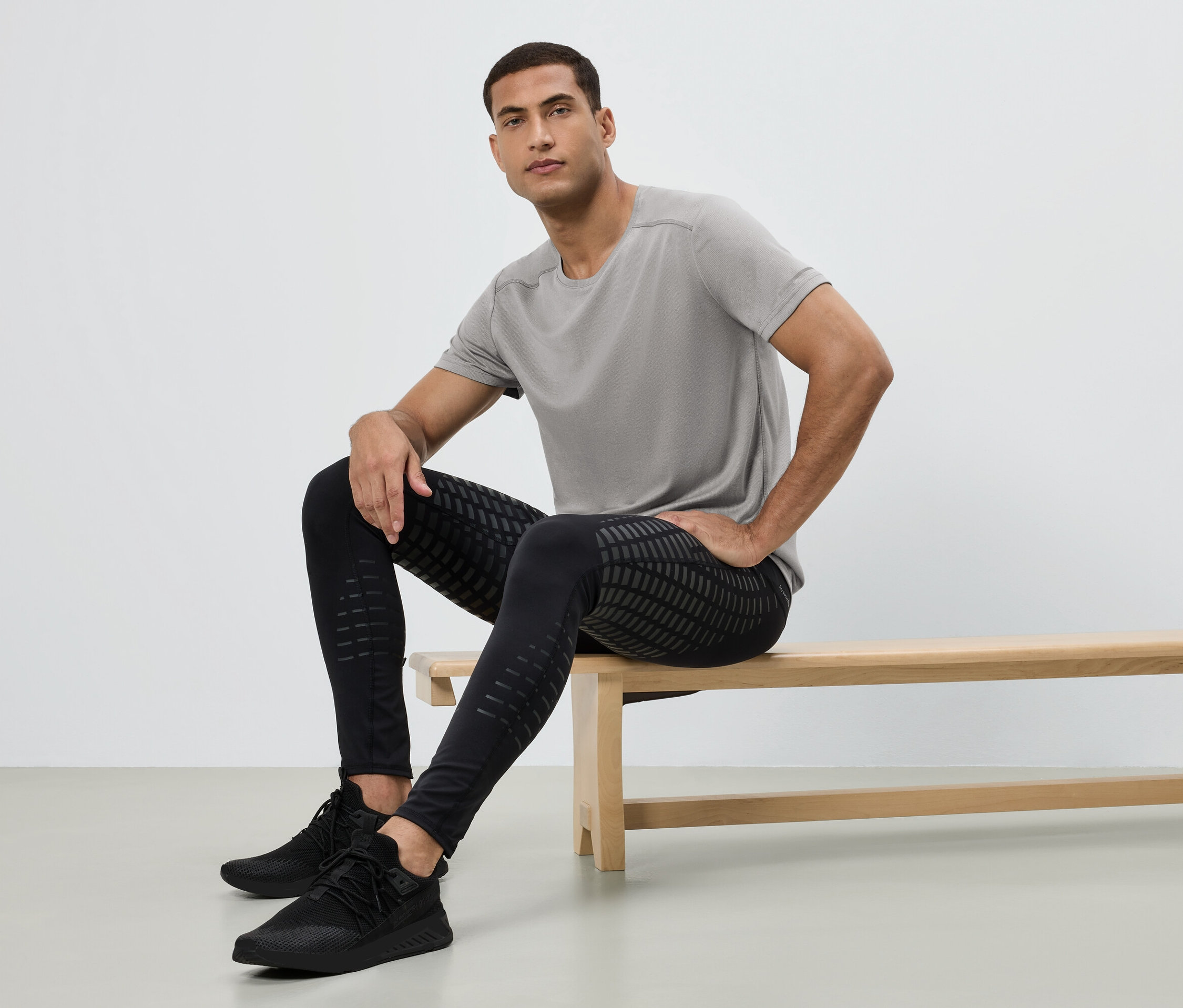 Muž sedí na lavičce, má na sobě šedé tričko, černé vzorované legíny a černé tenisky.