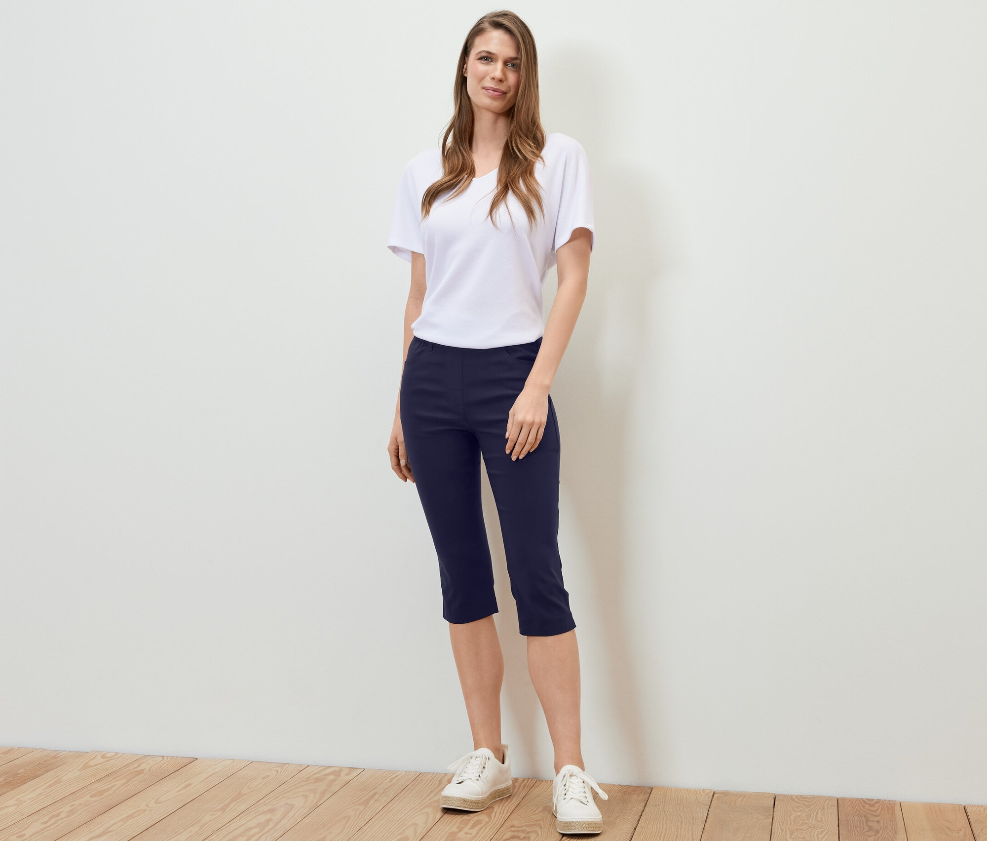 Žena stojí a má na sobě 3/4 bengalínové kalhoty tmavě modré barvy, bílé tričko a tenisky.