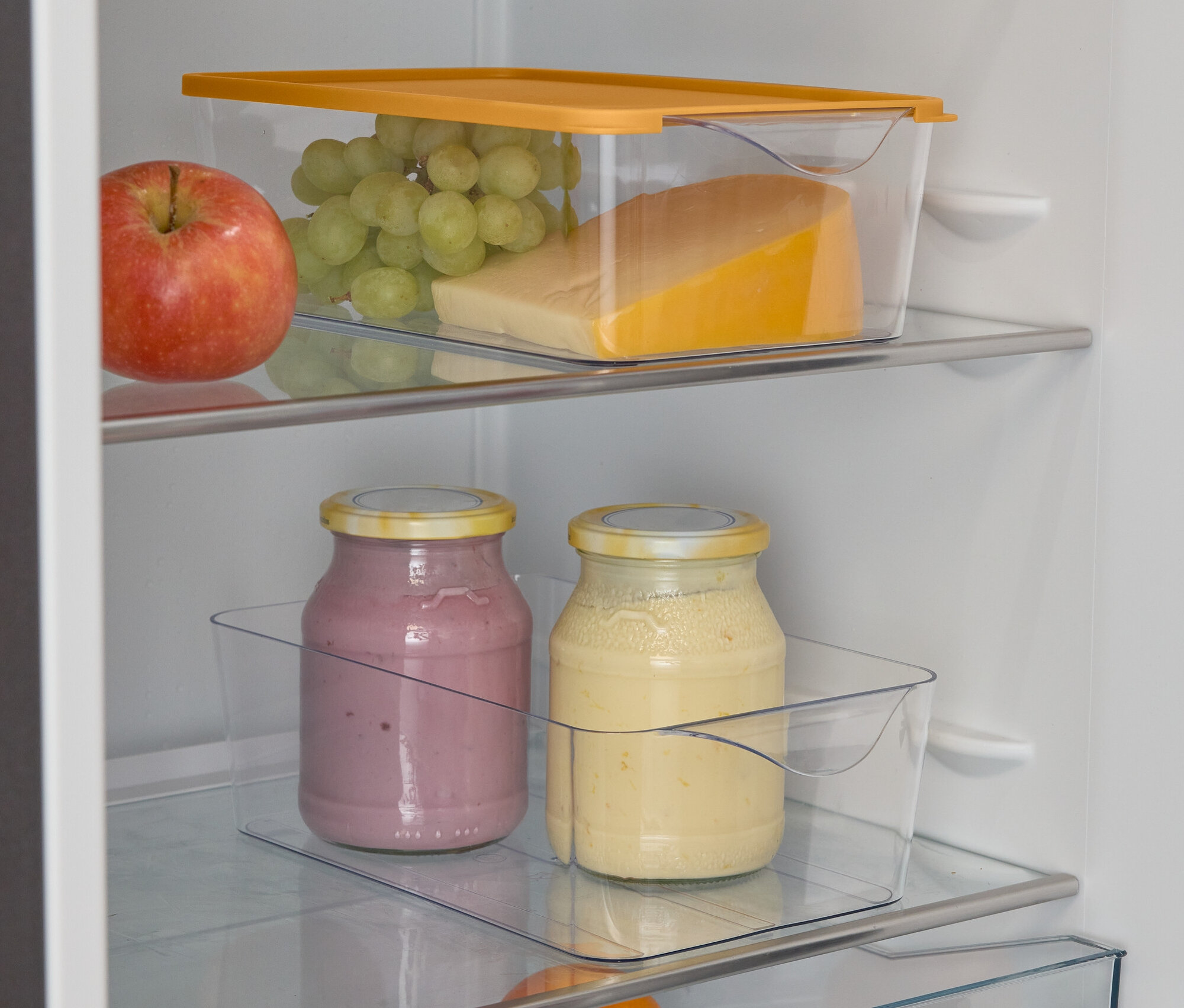 Otevřená lednice s jablkem, hrozny, sýrem, dvěma sklenicemi smoothie, pomerančem a limetkami.