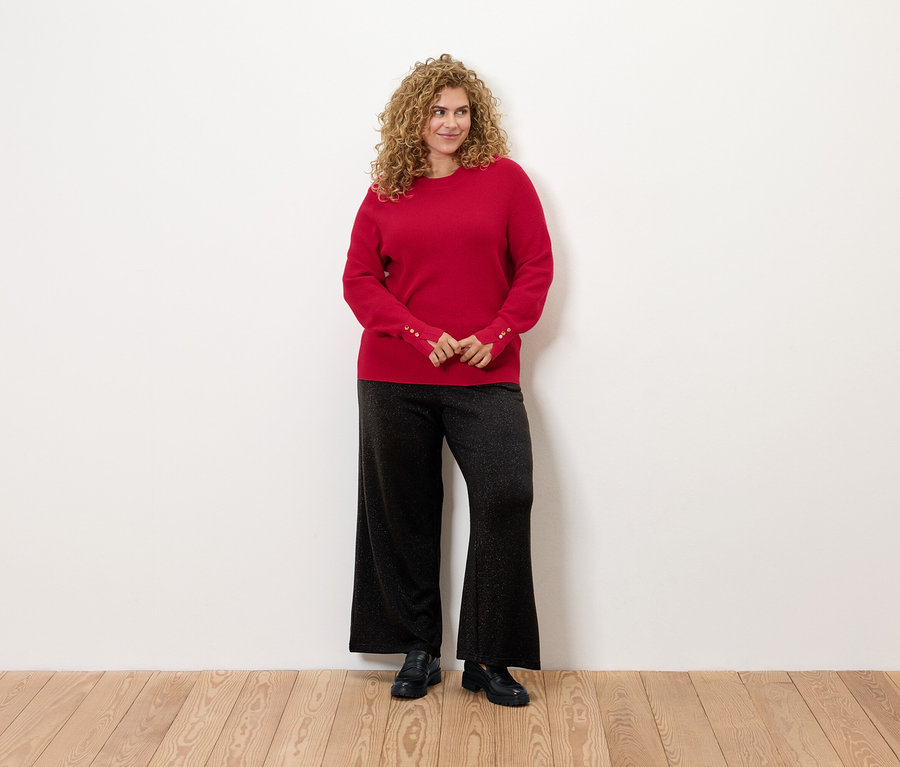 Žena s kudrnatými vlasy stojí a má na sobě červený svetr a černé kalhoty.