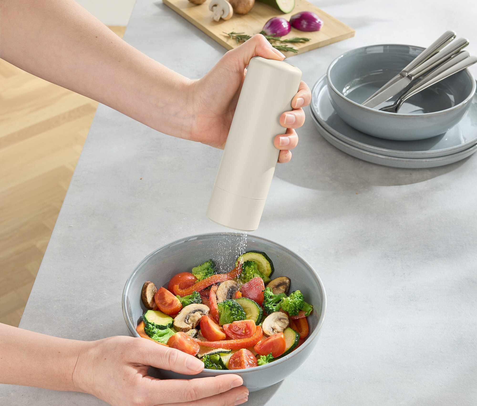 Osoba sype koření do mísy se salátem, na stole je prkénko s houbami, cibulí a cuketou.