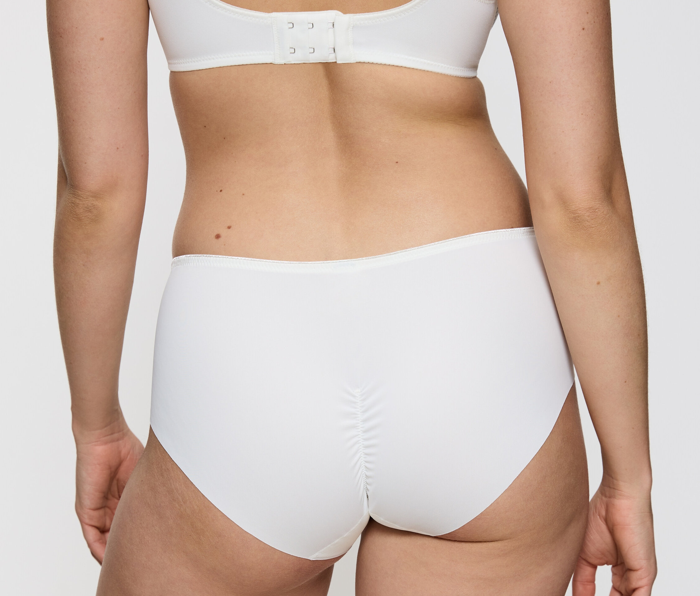 Zezadu je vidět žena, která má na sobě bílé kalhotky Triumph Essential »Minimizer Hipster X«, vanille a bílou podprsenku.