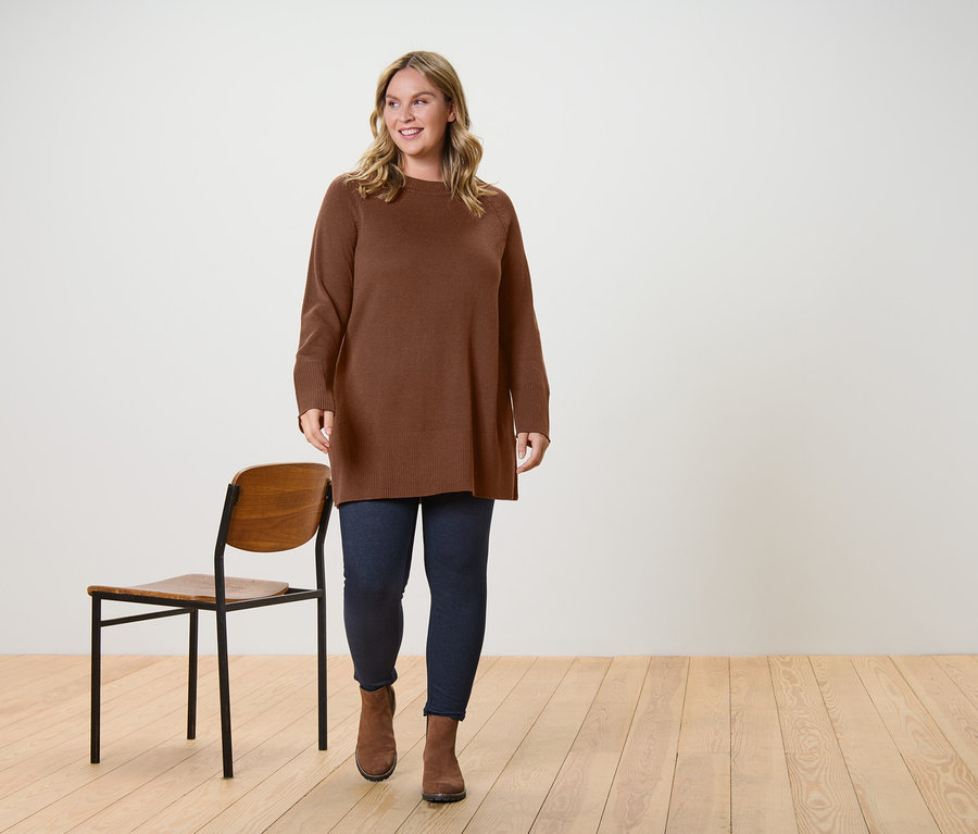 Žena kráčí a má na sobě dlouhý pletený svetr tmavě hnědé barvy a legíny v džínovém vzhledu s podšívkou.