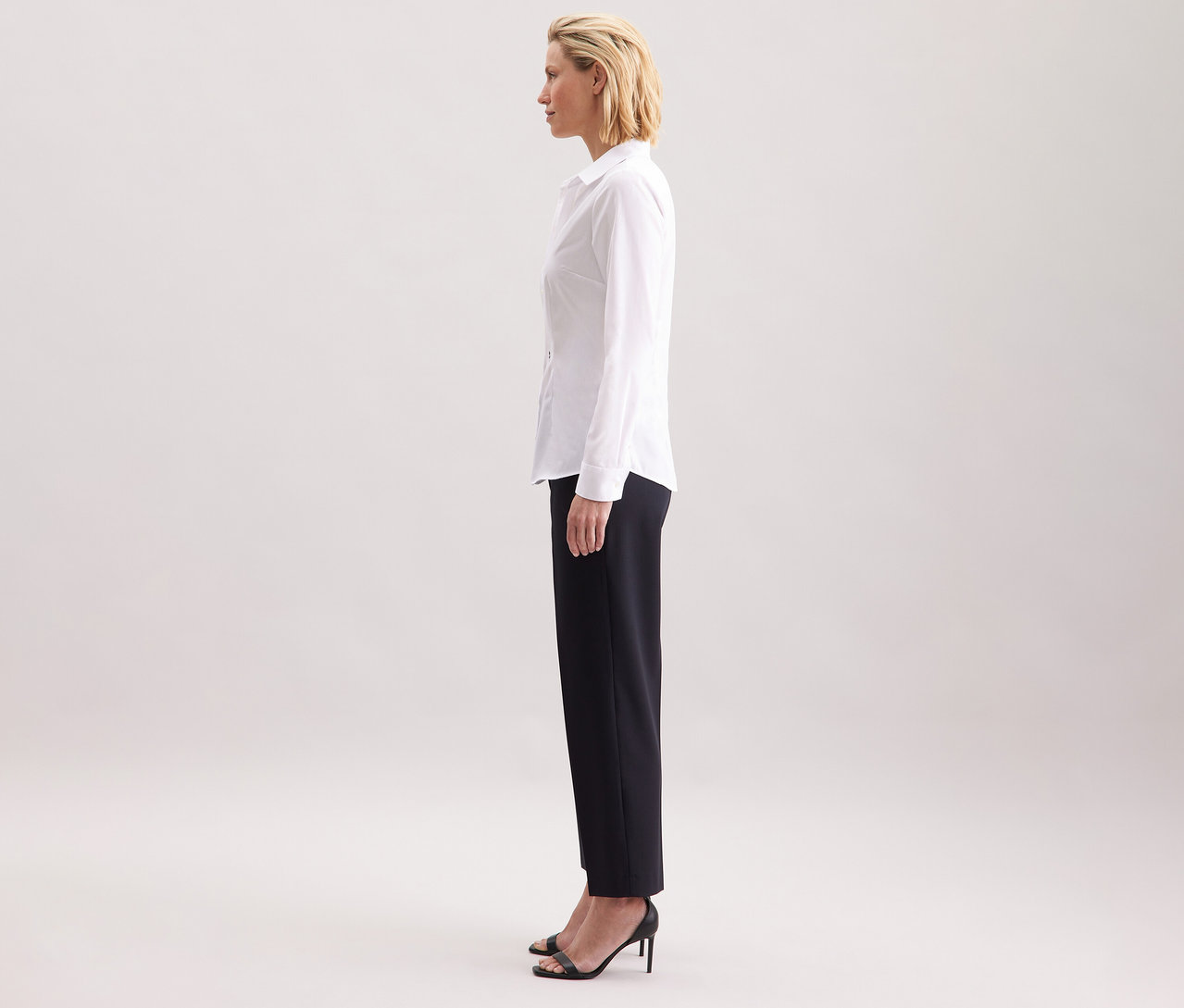 Žena v profilu nosí bílou halenku Seidensticker Slim Fit non iron, v kombinaci s tmavými kalhotami a černými vysokými podpatky.