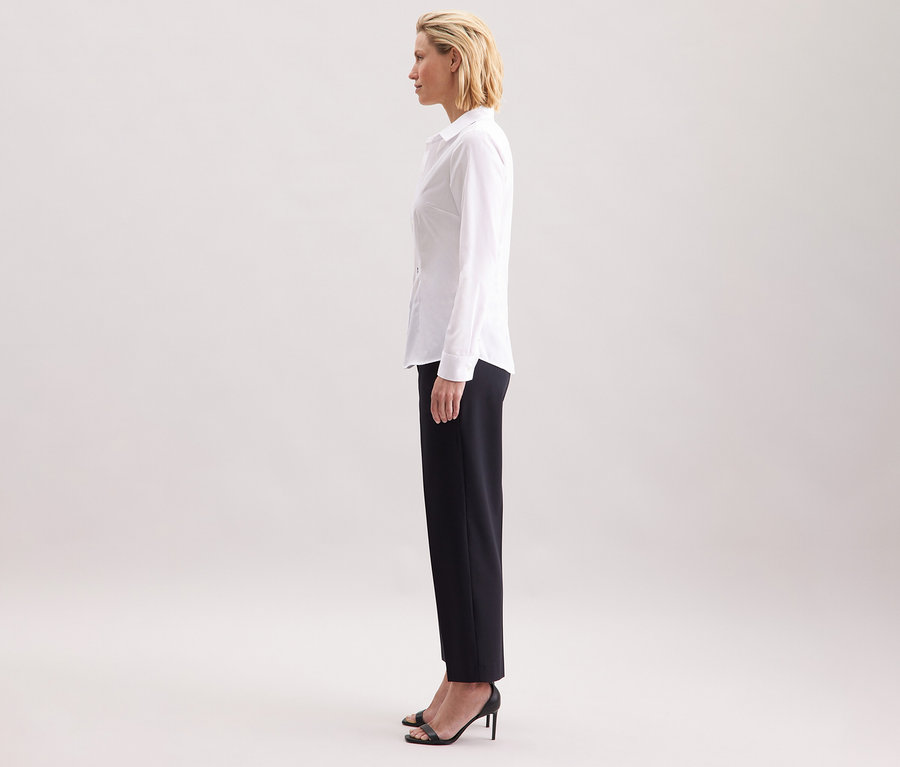 Žena v profilu nosí bílou halenku Seidensticker Slim Fit non iron, v kombinaci s tmavými kalhotami a černými vysokými podpatky.