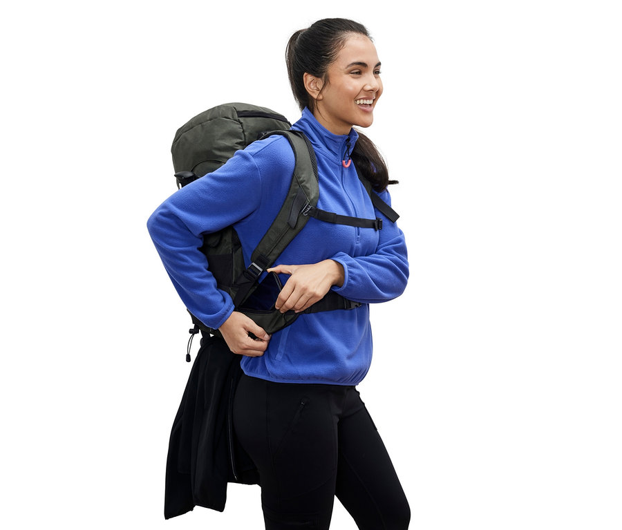 Usmívající se žena v modré fleecové bundě si upravuje popruh na zeleném turistickém batohu o objemu 40 l.