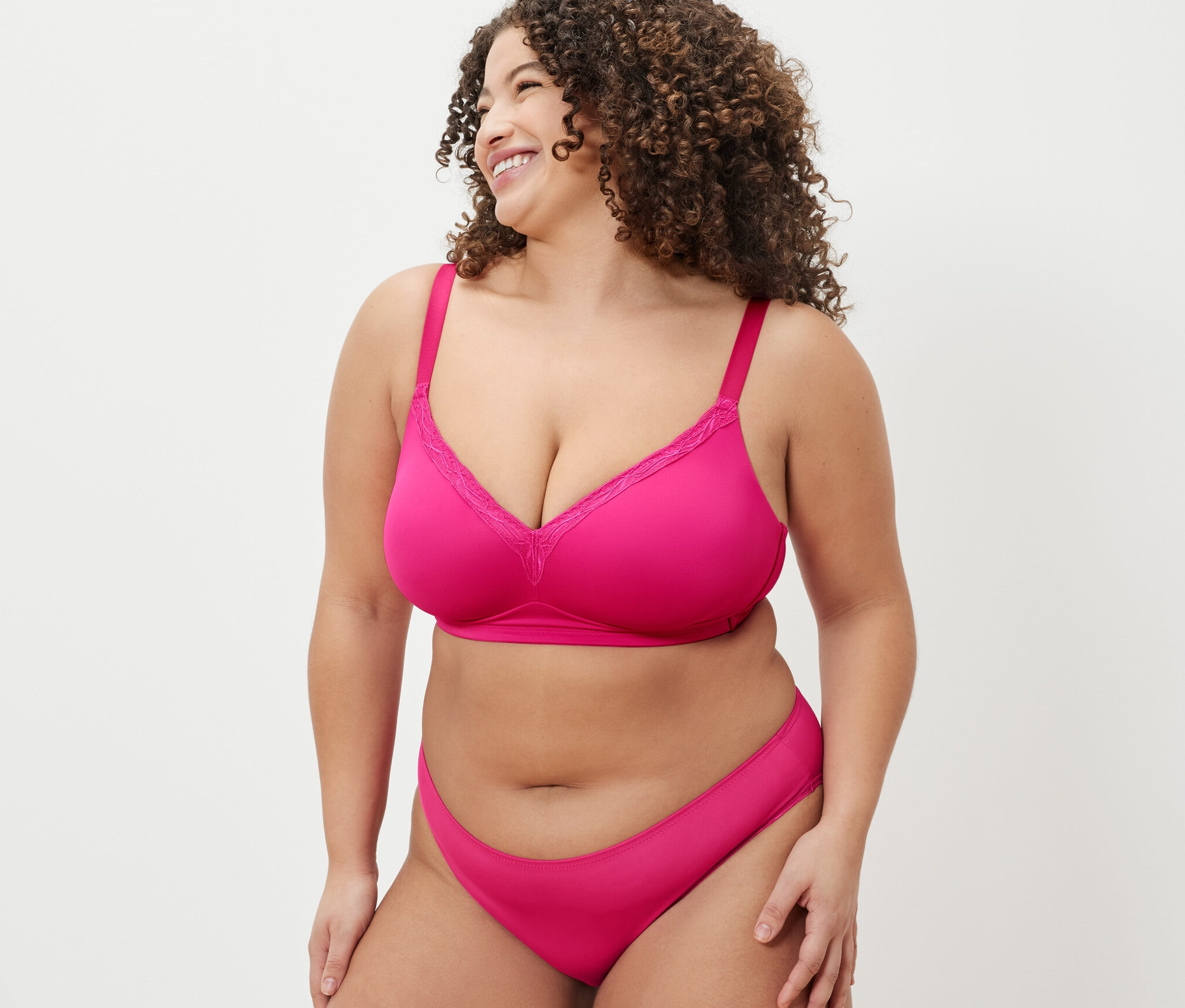 Žena v růžové soupravě spodního prádla s měkkou podprsenkou se dívá přes rameno a usmívá se.