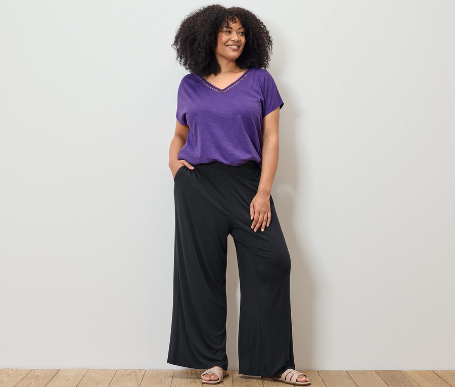 Žena pózuje v fialovém tričku z modalové směsi a černých kalhotách s širokými nohavicemi.