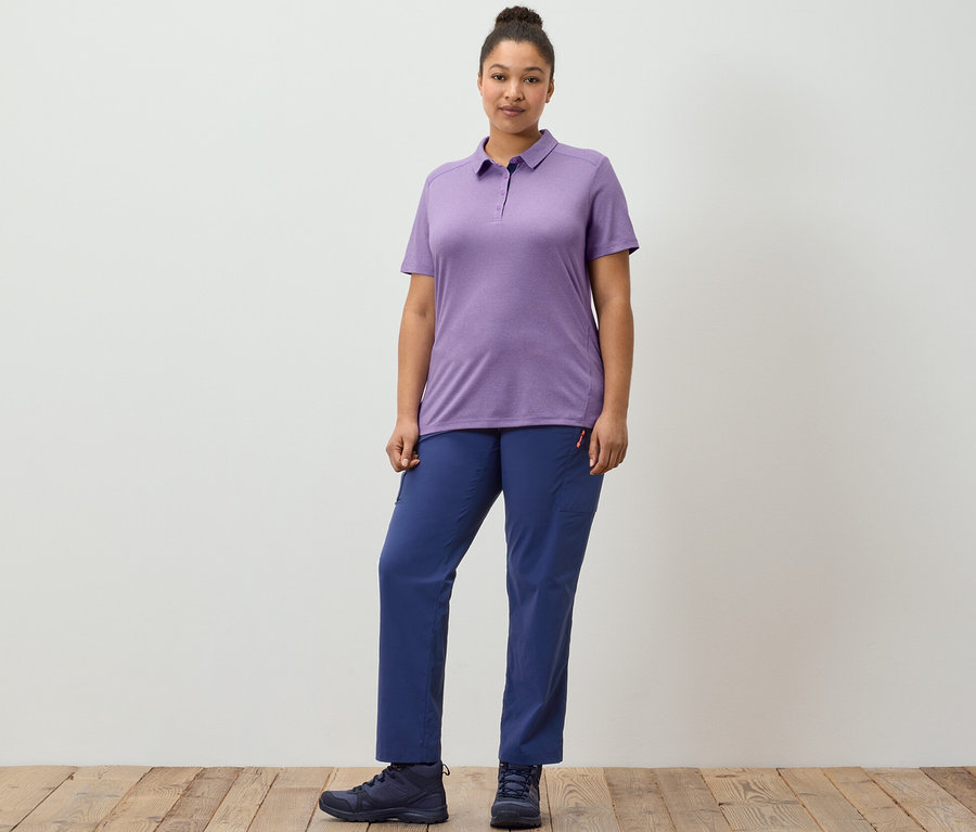 Žena pózuje na dřevěné podlaze v tmavě modrých outdoorových kalhotách a fialové funkční polokošili.