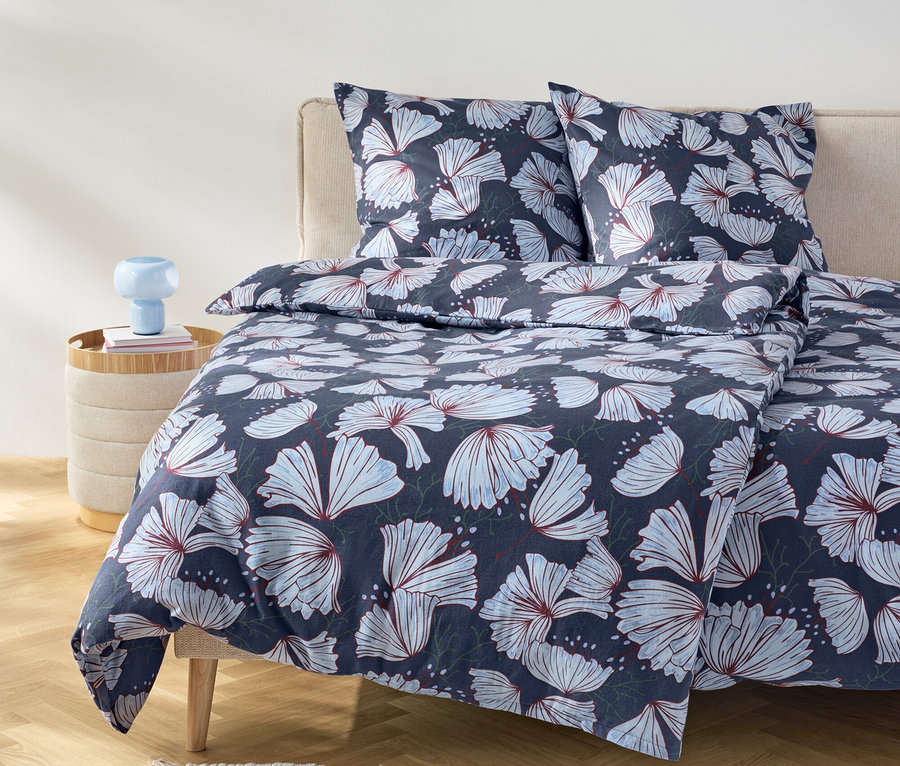 Postel s tmavě modrým povlečením s květinovým vzorem a dvěma polštáři na béžovém čele postele.