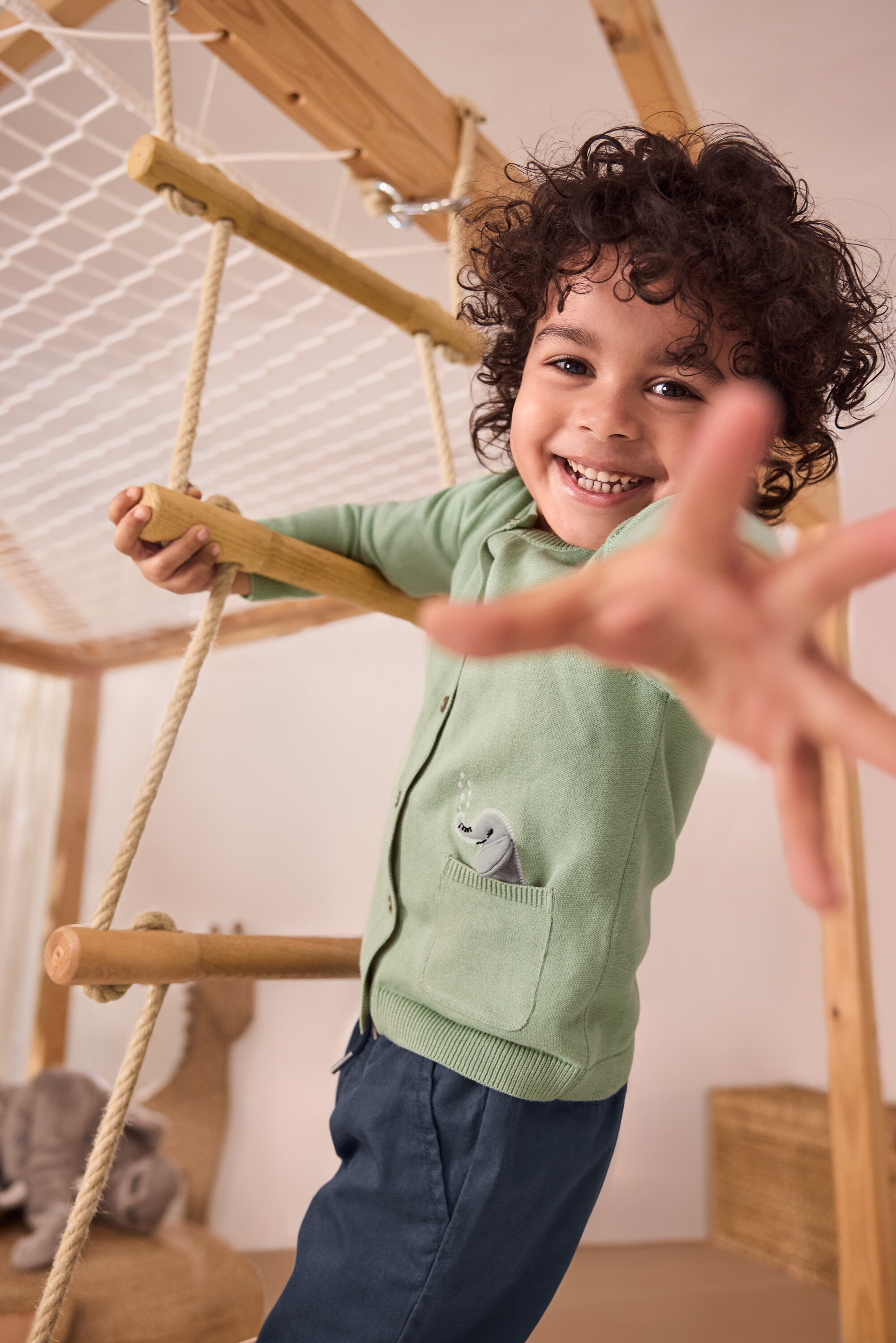 Usmívající se chlapec leze po provazovém žebříku. Má na sobě zelený dětský pletený svetr s aplikací a modré chlapecké navlékací kalhoty – Fit »Anton«.