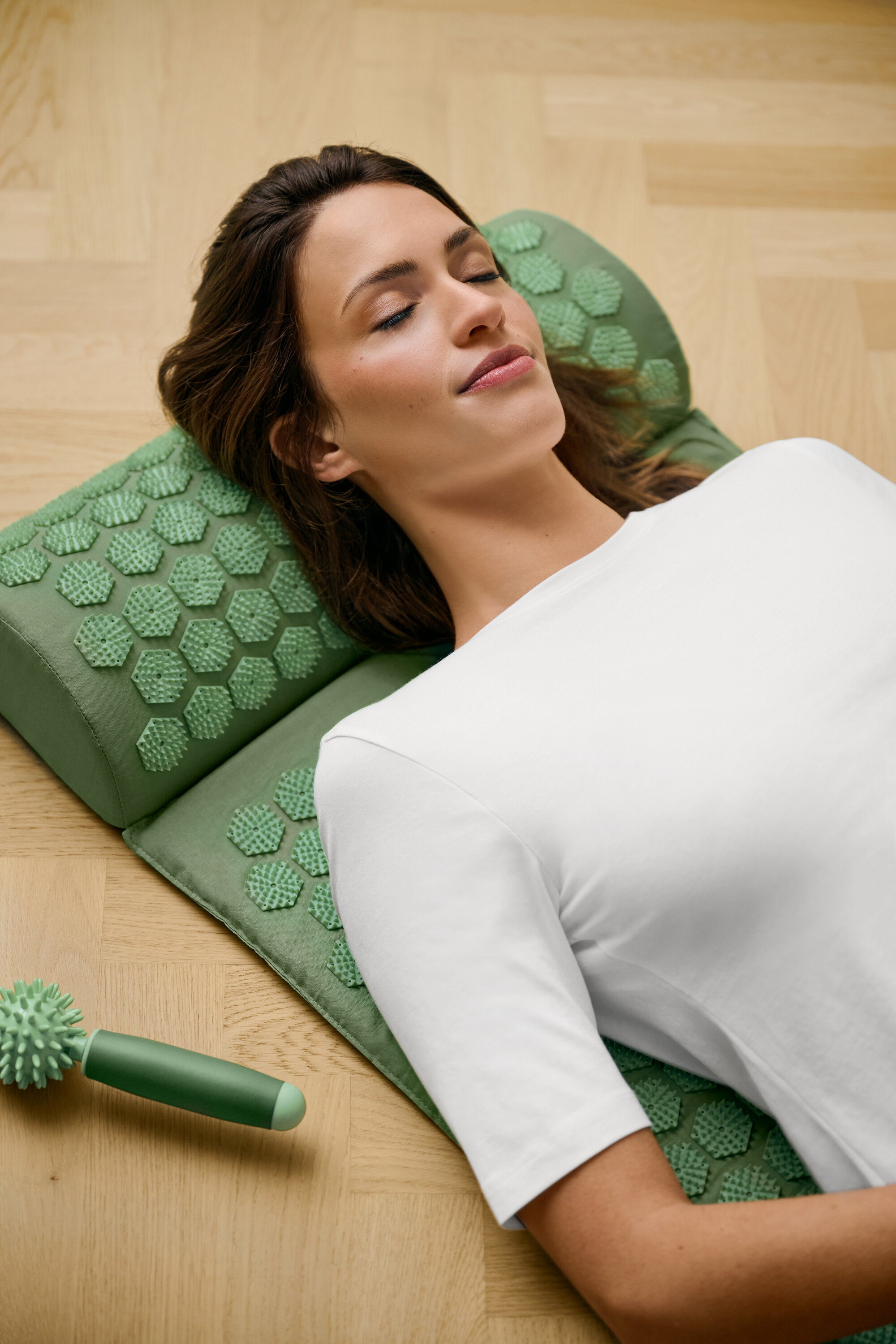 Žena leží na zelené akupresurní podložce a má na sobě bílé triko basic s polodlouhým rukávem.