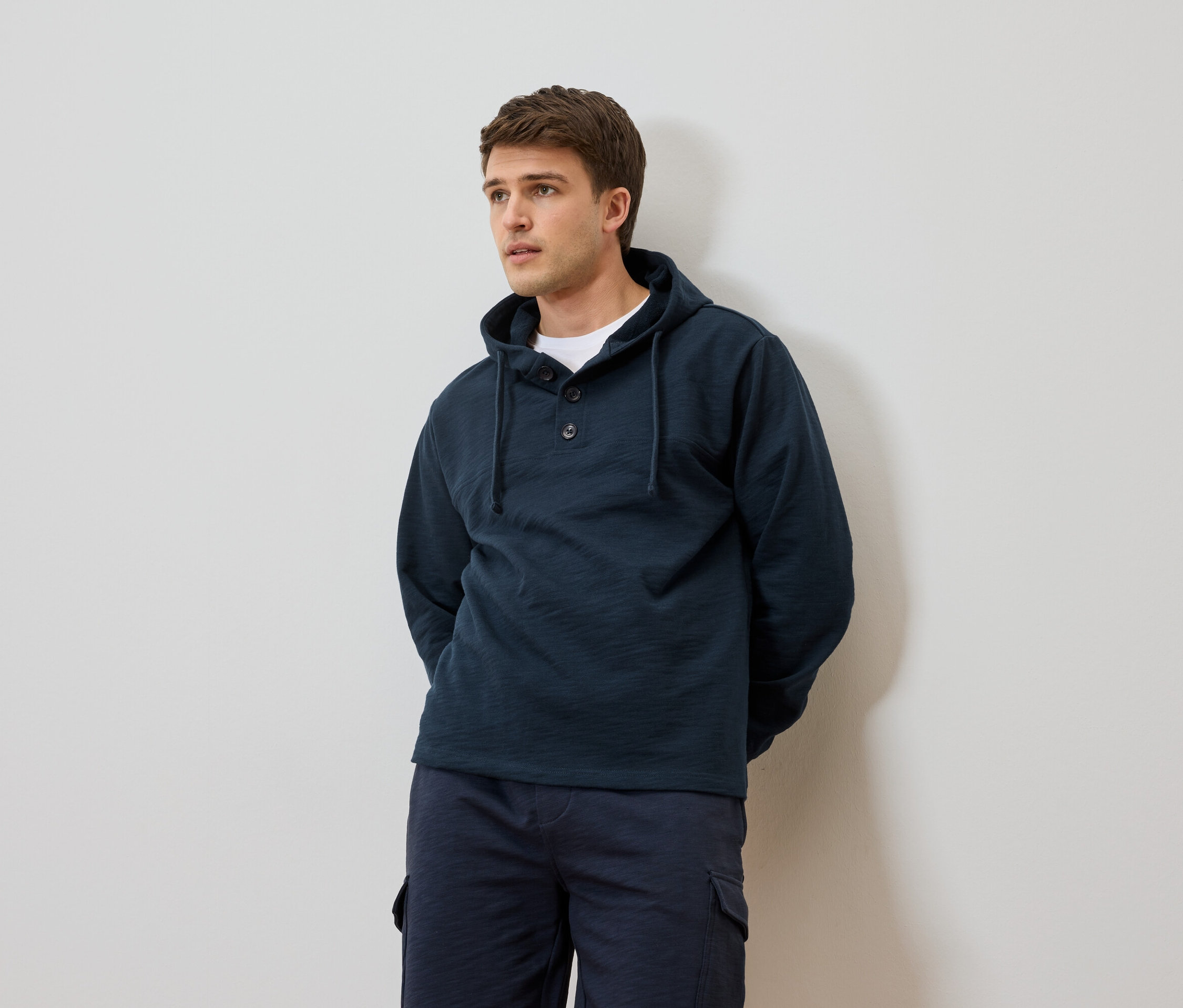 Muž v tmavě modré mikině s kapucí a šortkách před bílou zdí.