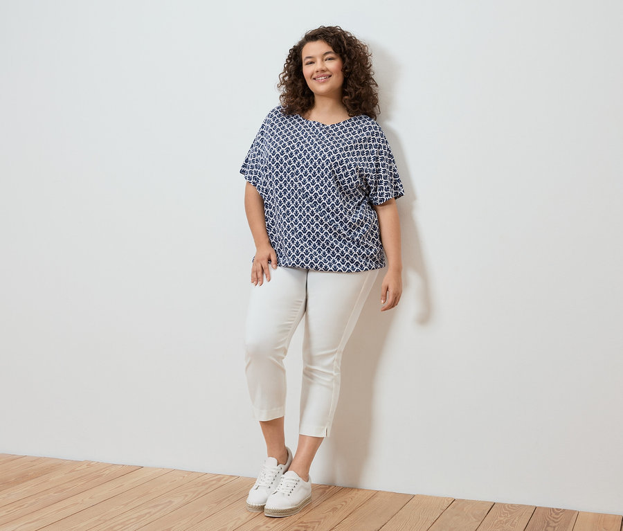 Žena s kudrnatými vlasy v modrobílé vzorované blůze, bílých kalhotách a bílých teniskách stojící před bílou zdí.