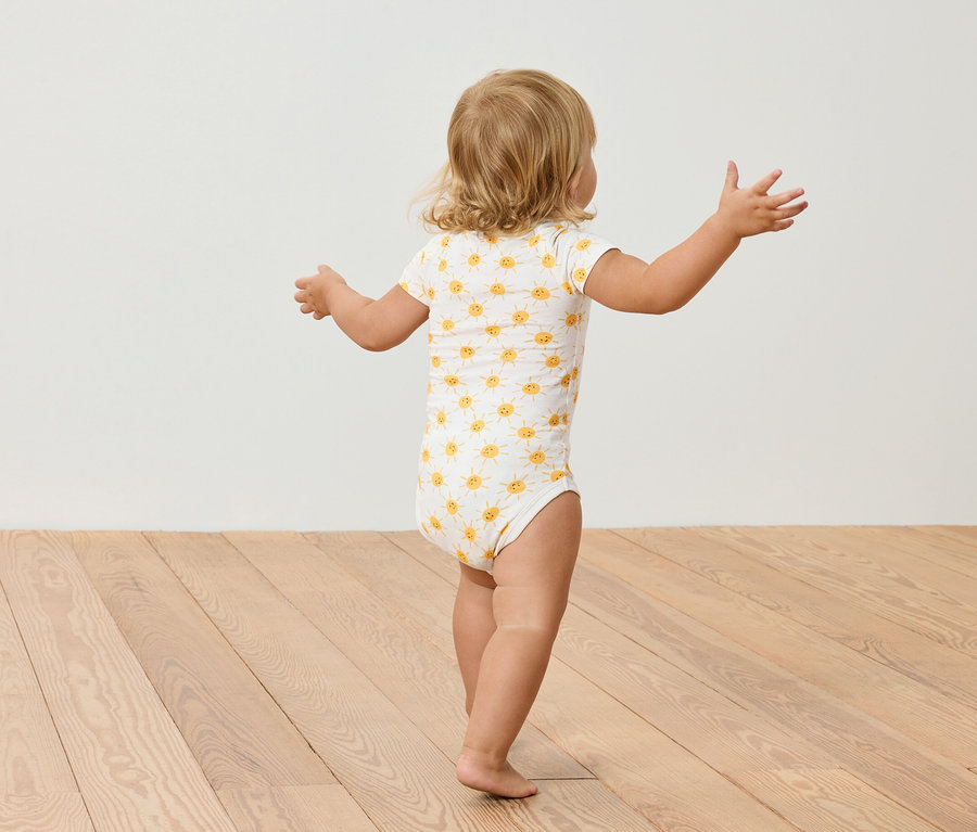 Malé dítě s blond vlasy v body s potiskem slunečnic stojí na dřevěné podlaze.