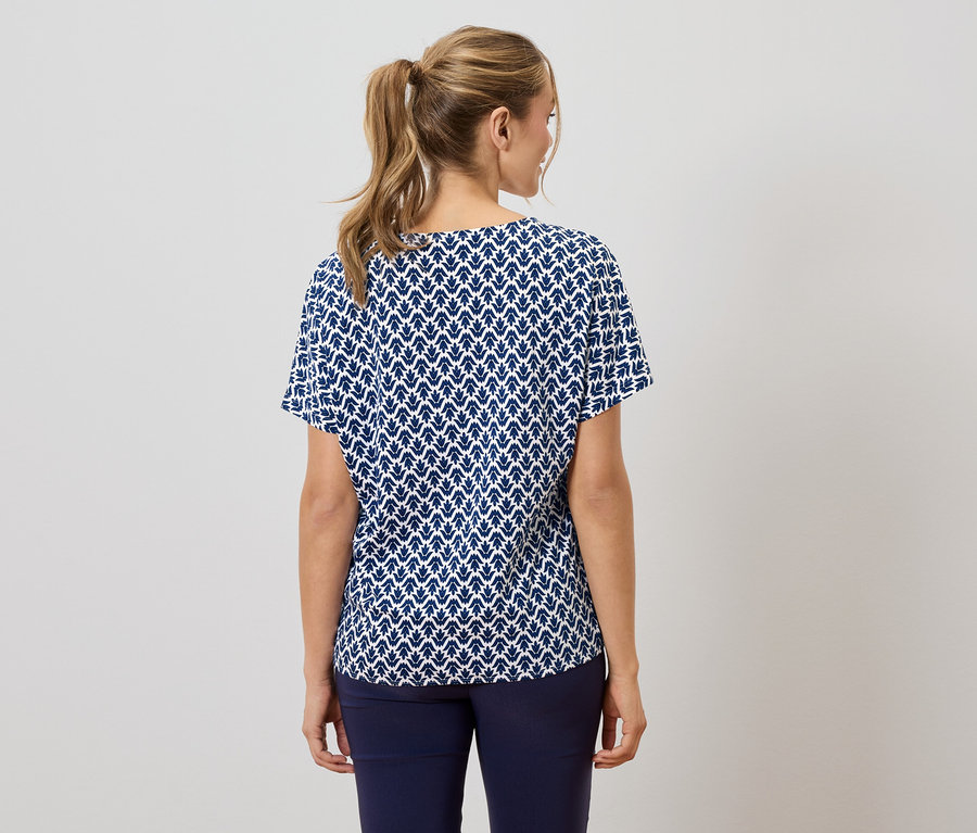 Pohled zezadu na ženu s blond copem, která má na sobě tmavě modrou a bílou halenku s krátkým rukávem s listovým vzorem a tmavě modré kalhoty.