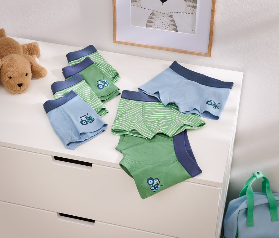 Fotografie světle modrých a zelených chlapeckých boxerek s motivem traktoru na komodě, vedle plyšového psa a modré cestovní tašky.