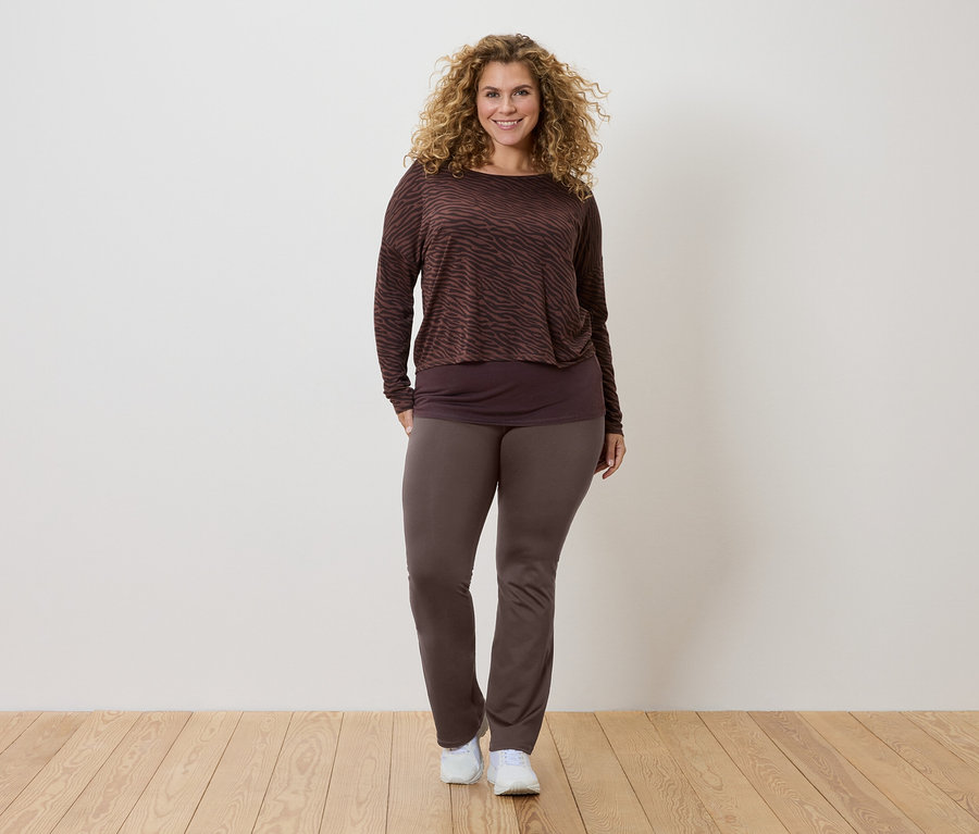 Žena s kudrnatými vlasy má na sobě hnědou vzorovanou halenku s dlouhým rukávem, tmavší top a hnědé kalhoty a bílé tenisky.