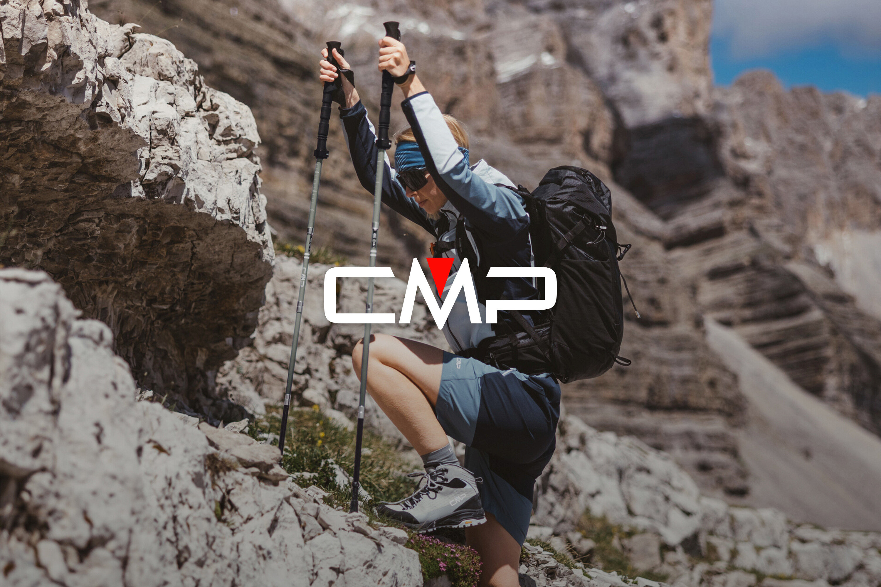 Žena s batohem a trekovými holemi leze po skalách. Má na sobě trekové boty, šortky a bundu.