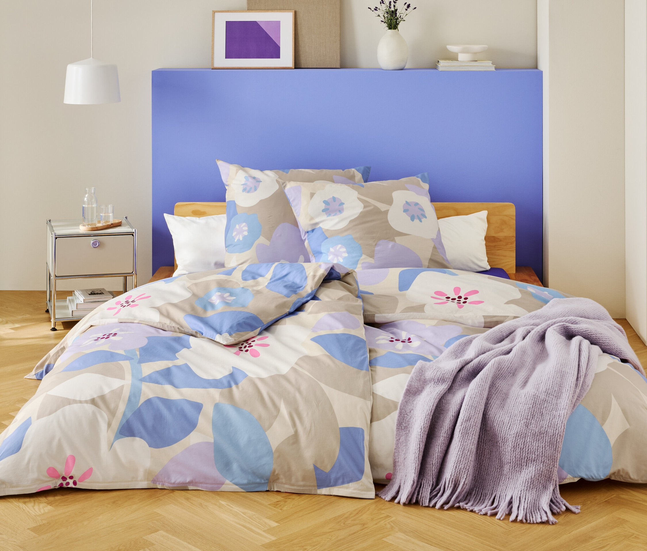 Ložnice s postelí přikrytou květinovovým ložním prádlem a fialovou dekou.