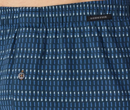 Detail modré látky se vzorem světlých a tmavých modrých obdélníků, nahoře elastický pas, černý štítek s nápisem „SCHIESSER“ a knoflík s nápisem „SCHIESSER EST. 1875“.