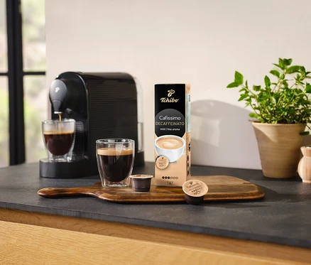 Kávovar nalévá kávu do sklenice vedle balení kapslí Cafissimo Decaffeinato.