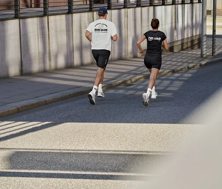Muž v bílém sportovním tričku s nápisem „RUN CLUB“, černých sportovních šortkách a tenisových ponožkách Coffee Crew a žena v černém sportovním tričku s nápisem „RUN CLUB“ a krátkých sportovních legínách ActivePower běží po silnici.