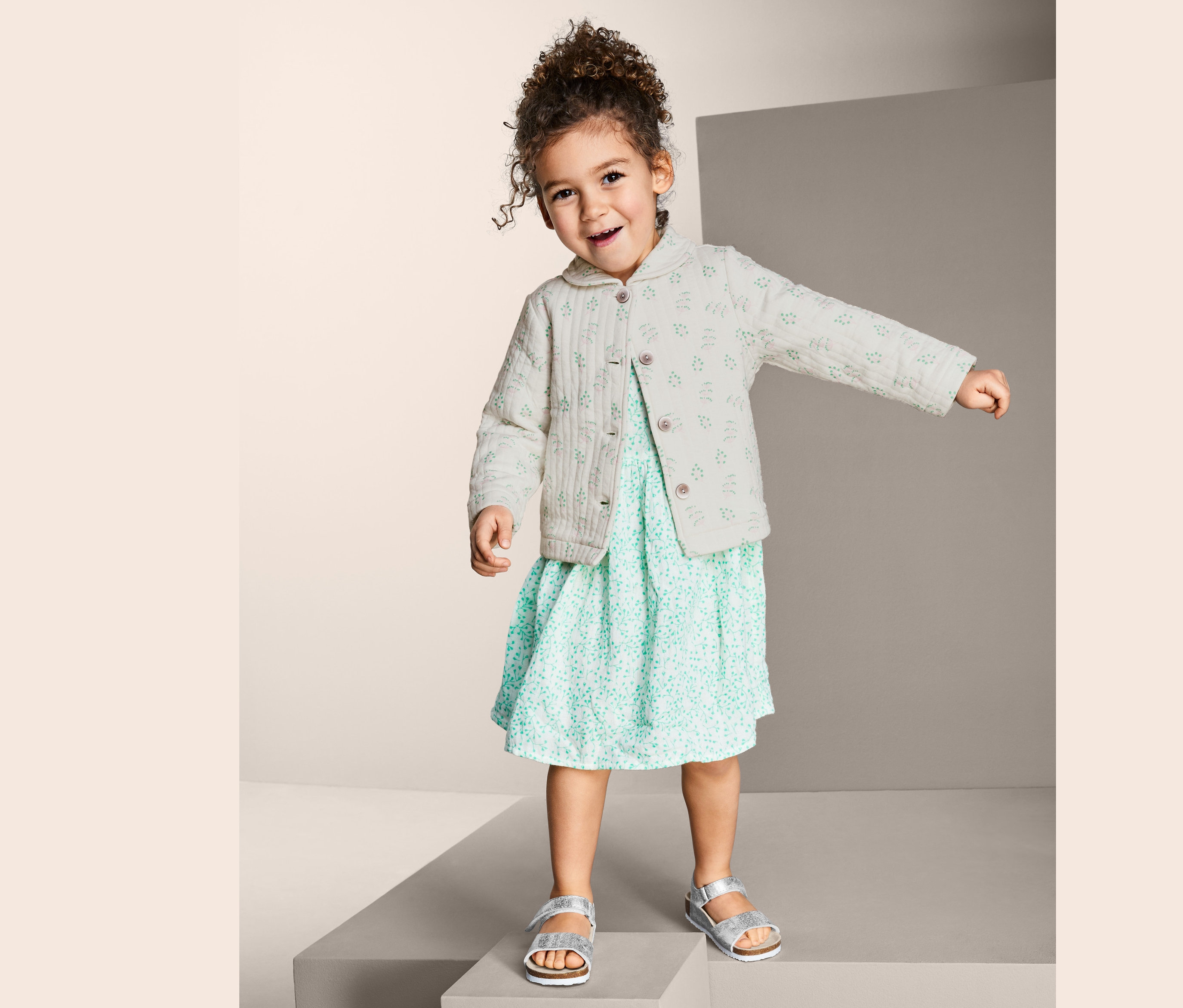 Holčička stojí na podstavci a má na sobě prošívaný bavlněný kabátek a letní šaty.