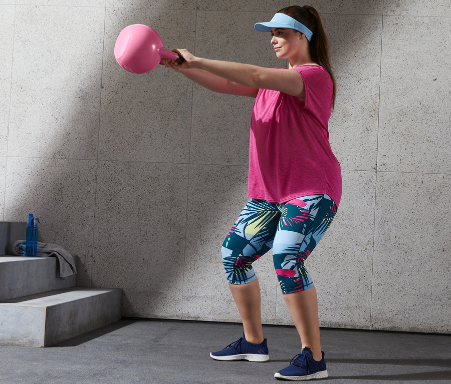 Žena cvičí s růžovou kettlebell v růžovém sportovním tričku a 3/4 sportovních legínách.