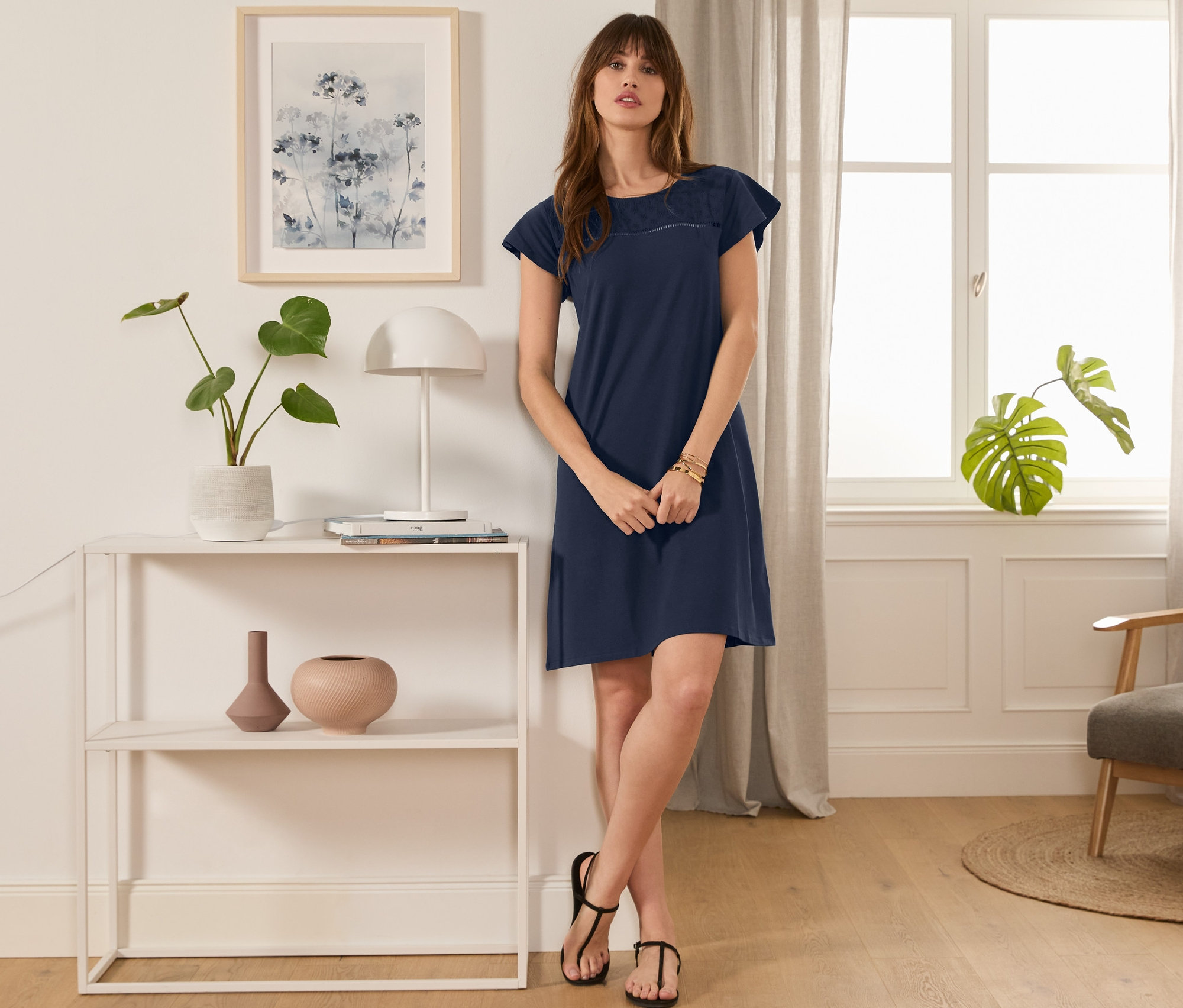 Žena stojí v místnosti a má na sobě tmavě modré žerzejové šaty s ažurovou výšivkou.