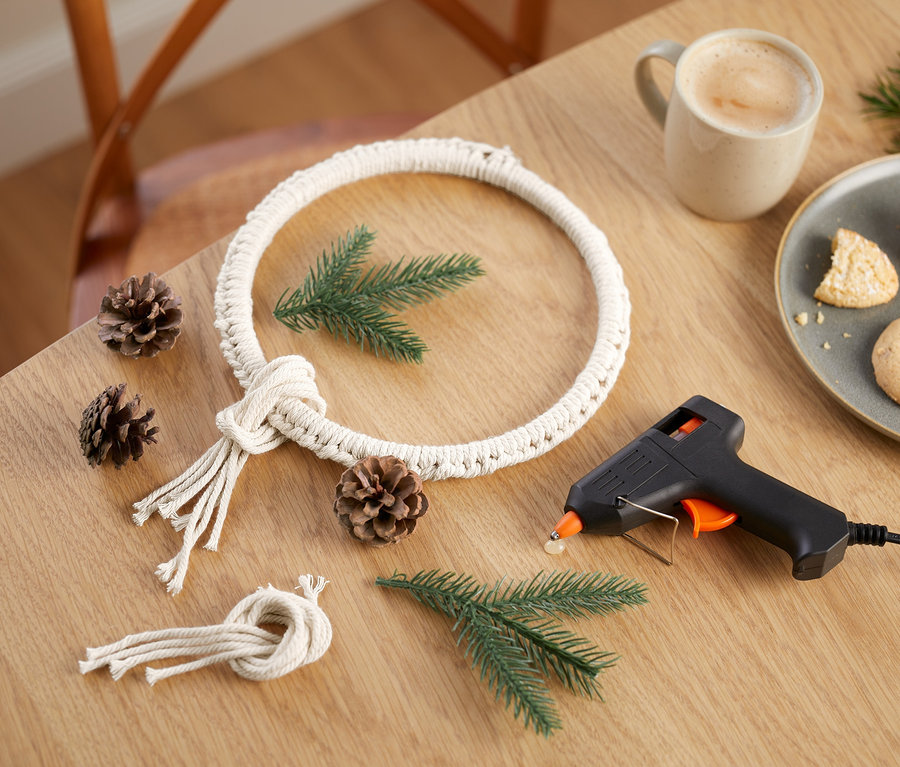 Na stole leží sada na výrobu drhaného zimního věnce, šišky, větvičky, pistole na tavné lepidlo, hrnek s kávou a talíř s sušenkami.