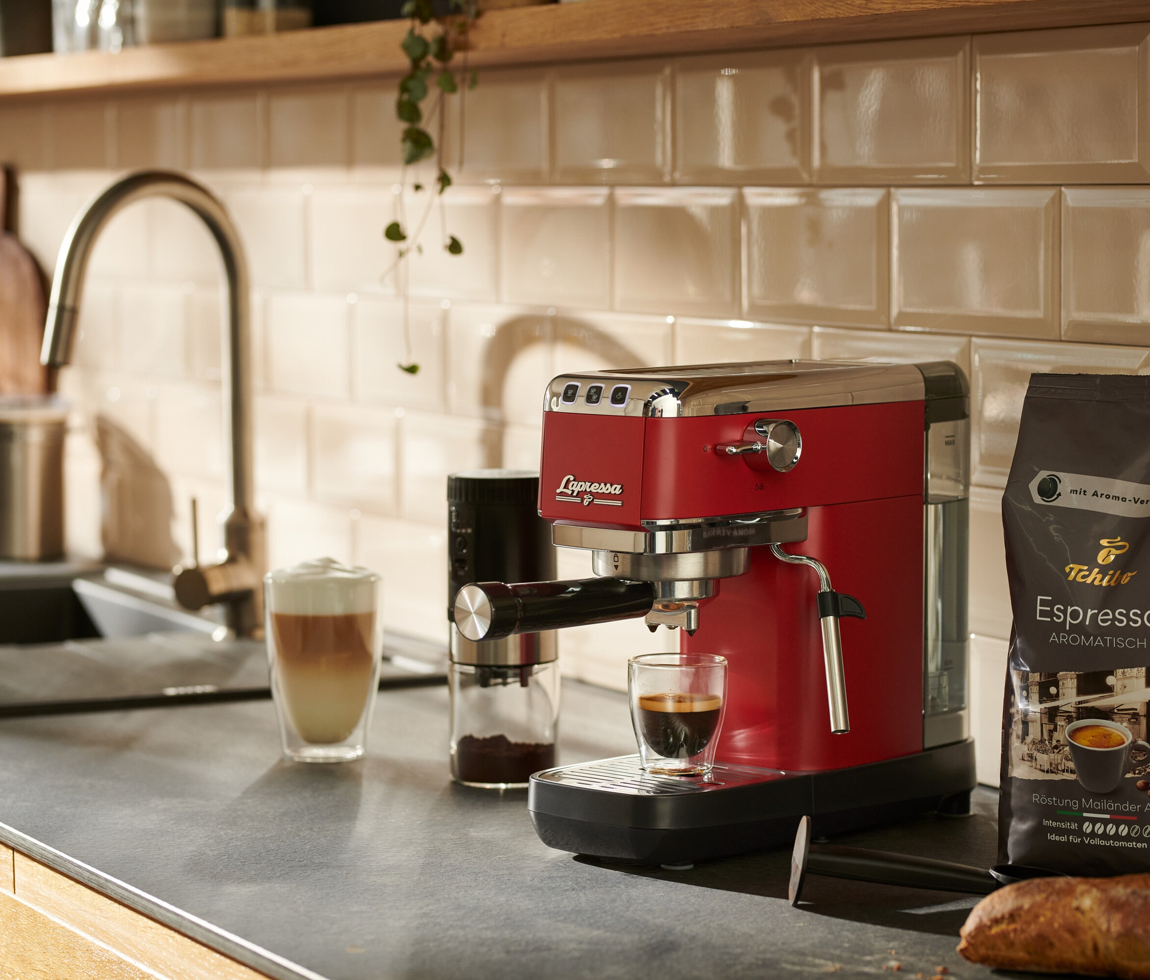 Červený pákový kávovar »Lapressa« stojí na kuchyňské lince vedle sklenice s kávou.