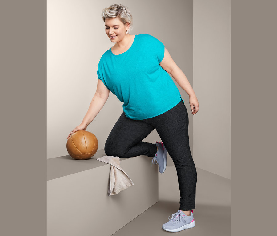 Žena v tyrkysovém sportovním tričku a sportovních kalhotách se opírá o hnědý medicinbal.