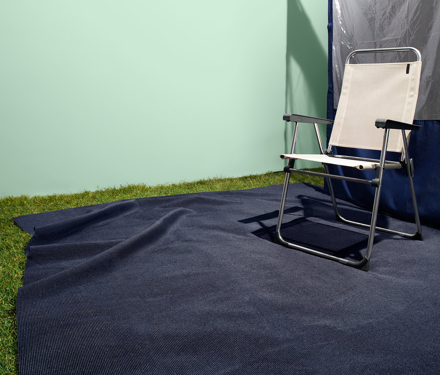 Tmavě modrý koberec na stanování leží na trávě vedle skládací židle a stanu.