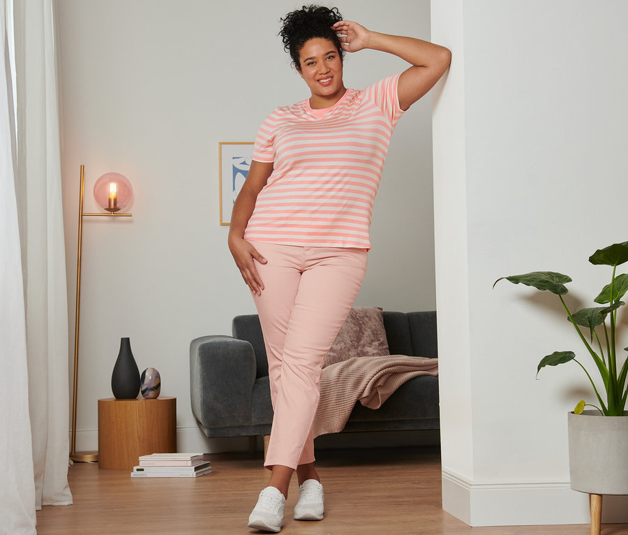 Žena pózuje opřená o zeď v růžovém proužkovaném tričku, světle růžových džínách slim fit a velurových teniskách.