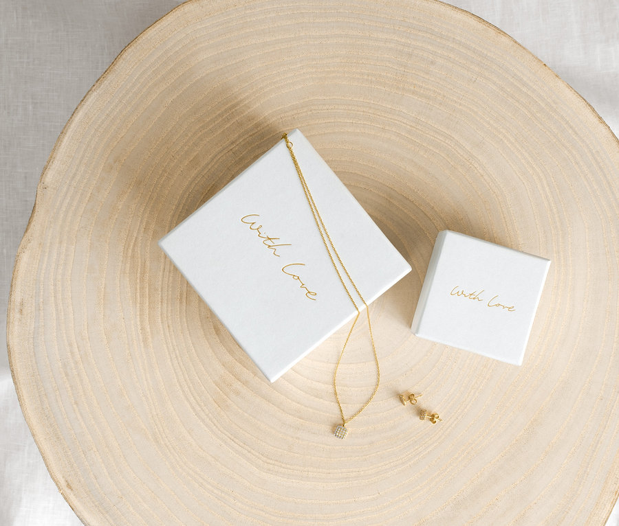 Zlatý řetízek se zirkony Cushion a zlaté náušnice se zirkony Cushion na dřevěném podkladu vedle bílých krabiček.