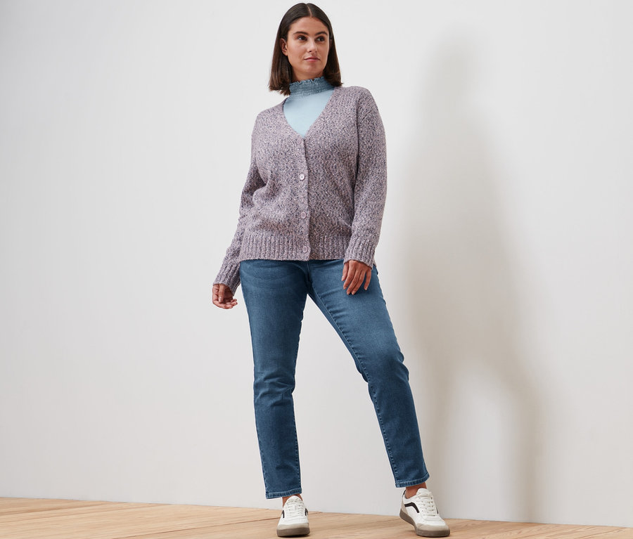 Žena pózuje v halénkovém triku se stojáčkem kouřově červené barvy, džínách slim »Fit Emma« středně modré barvy, pleteném kabátku a kožených teniskách.