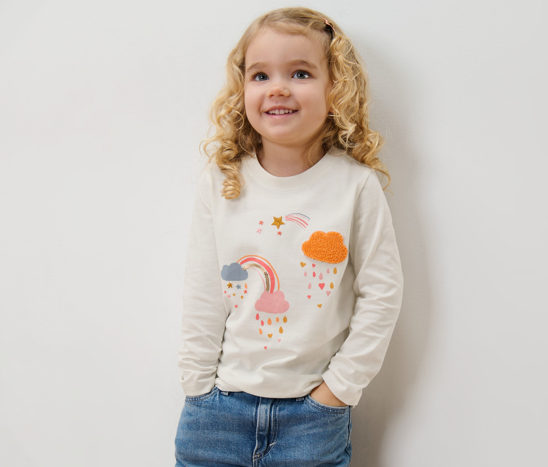 Usmívající se dívka pózuje v džínách a tričku s dlouhým rukávem s potiskem duhy.