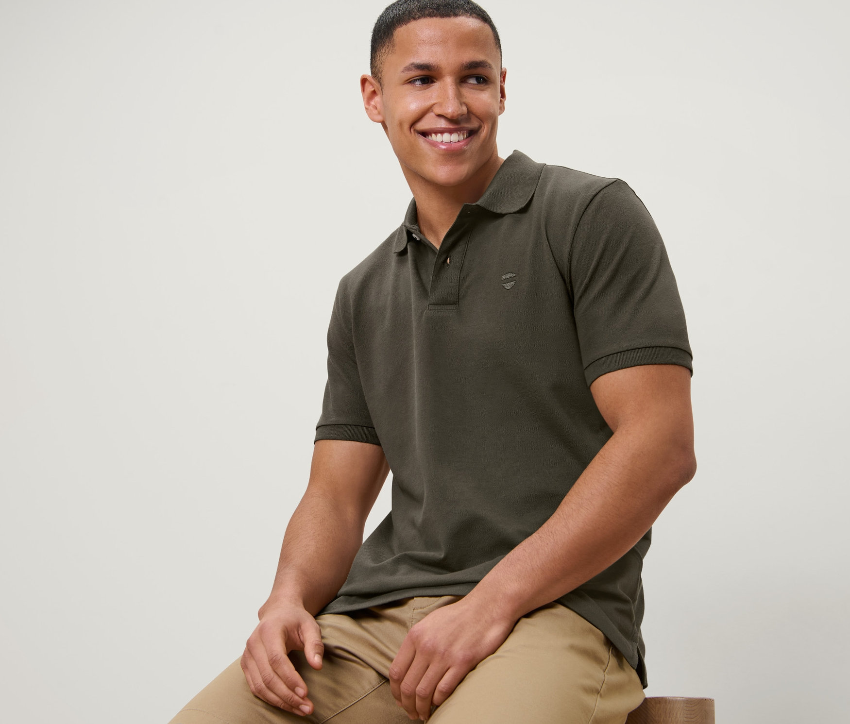 Muž sedí na stoličce a má na sobě olivovou polokošili z piké a béžové šortky.