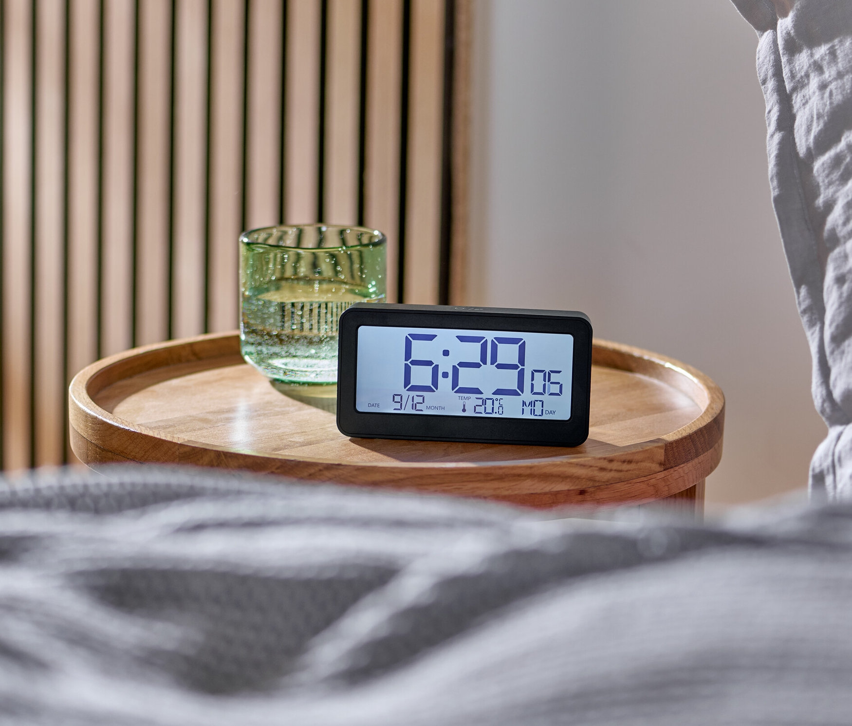 Digitální LCD budík řízený rádiovým signálem na dřevěném stolku vedle postele.