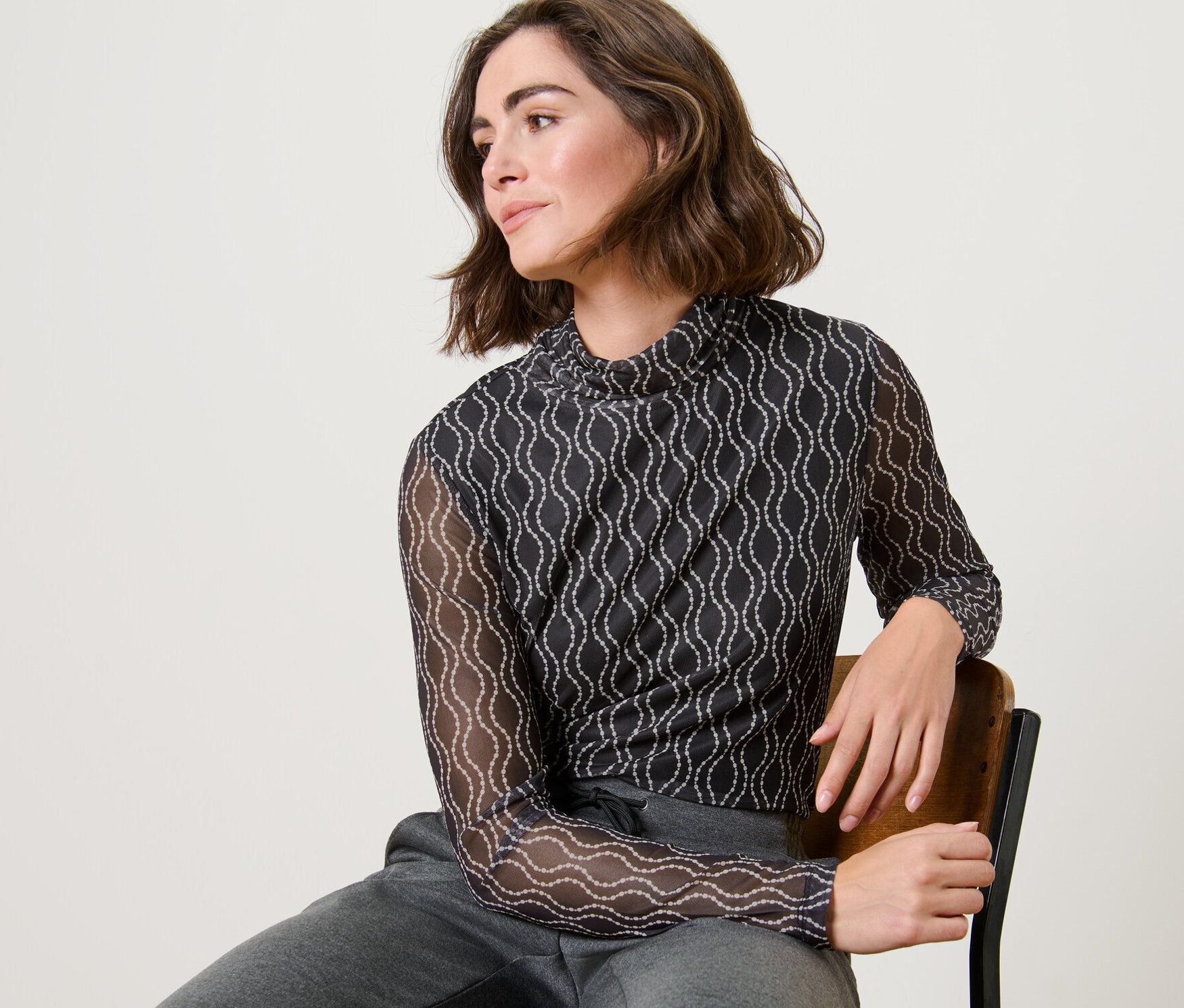 Žena sedí na židli a má na sobě síťované rolákové triko s potiskem a šedé tepláky.