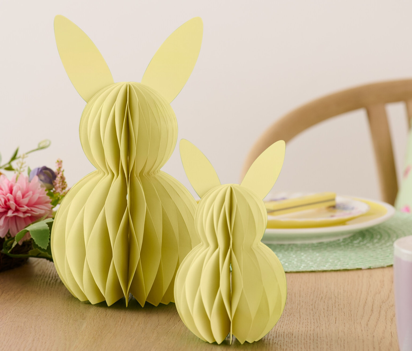 Dva papíroví velikonoční zajíčci stojí na stole vedle květin a talířů.